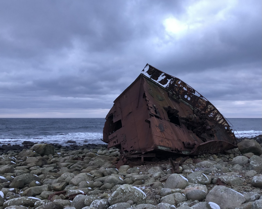 Skipsvrak på steinstrand med mørke skyer bak.