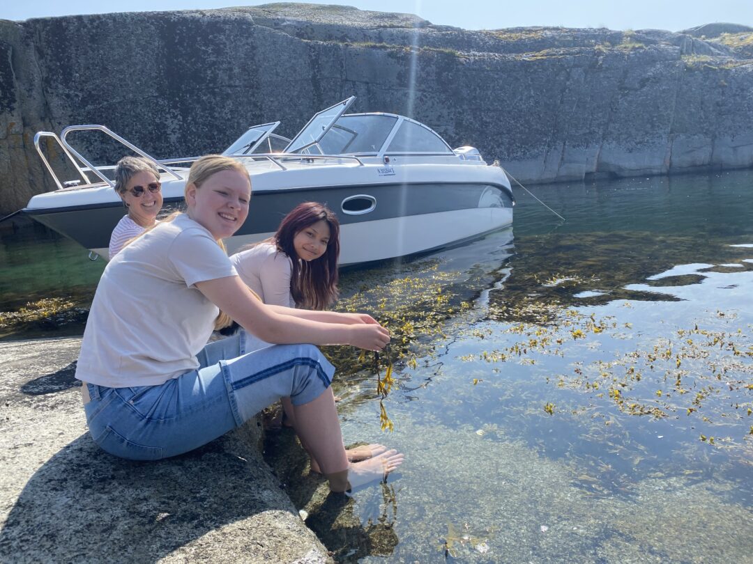 Jenter og lærer i strandkanten foran en båt.