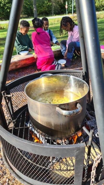 Bilde av grønnsakssuppe i kjele på bålpanne, med fire elever i bakgrunnen.