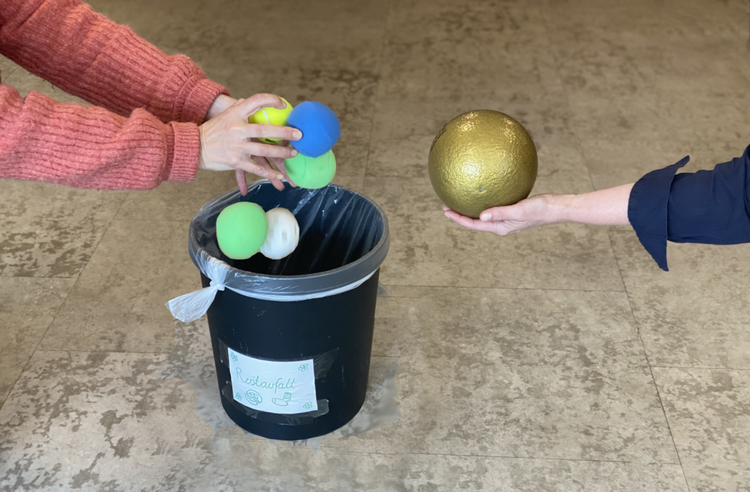 Fem små baller slippes i en søppelbøtte. En større gullfarget ball rekkes fram.
