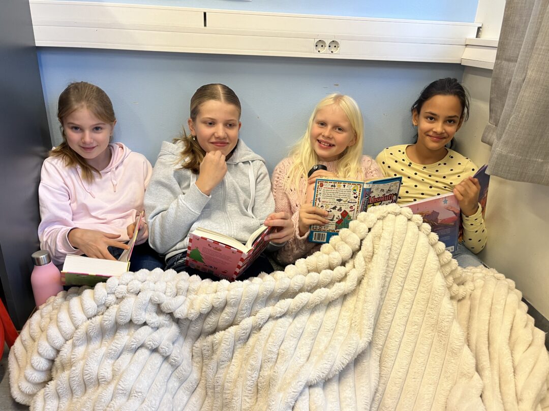 Bilde av fire jenter som sitter på gulvet og leser, med et teppe over seg.